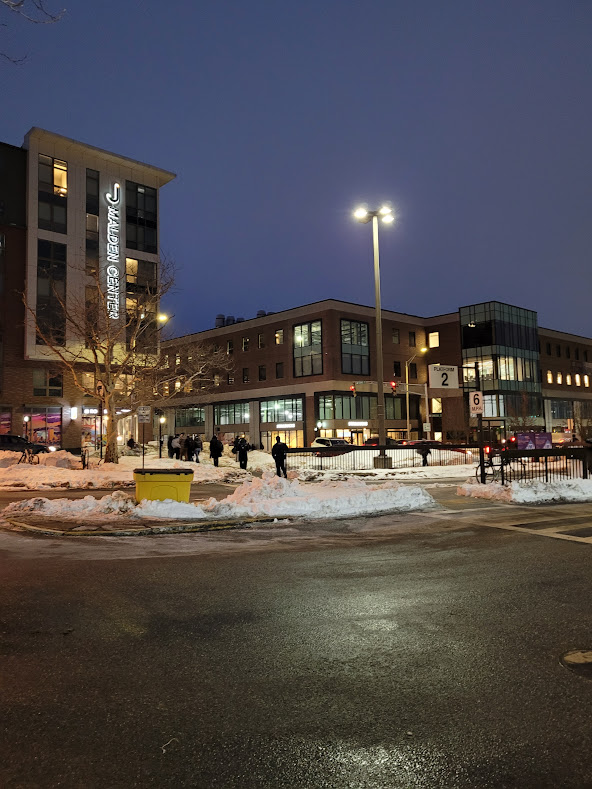 A wide, clear sidewalk in downtown Malden Center leads commuters toward the MBTA station, with snow piled at the curb.