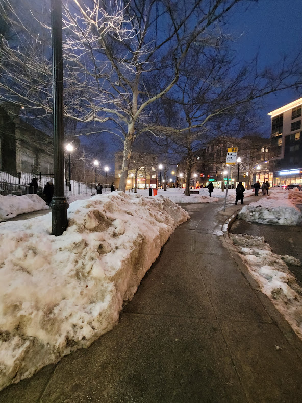 A wide, clear sidewalk in downtown Malden Center leads commuters toward the MBTA station, with snow piled at the curb.