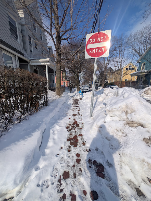 Brick sidewalk partially cleared in Porter Square. A red “Do Not Enter” sign and triple-decker homes along the street are visible.