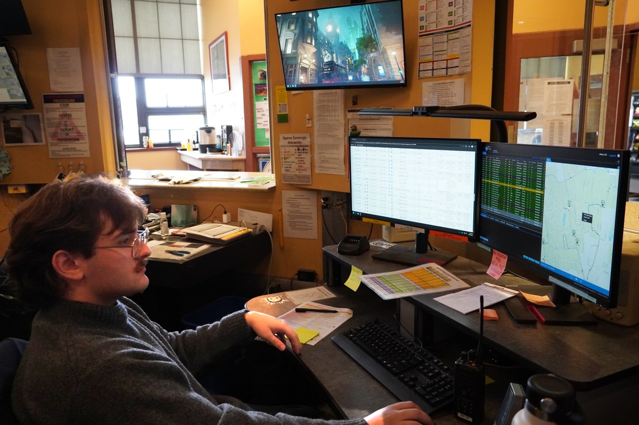 A young man with a mustache and glasses sits in front of two computer screens in a cluttered office.