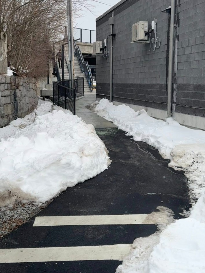 A shovelled paved path runs through the snow between a fence and a small cinderblock building. In the distance beyond the building is a train platform and a stairway leading up to a street overpass.