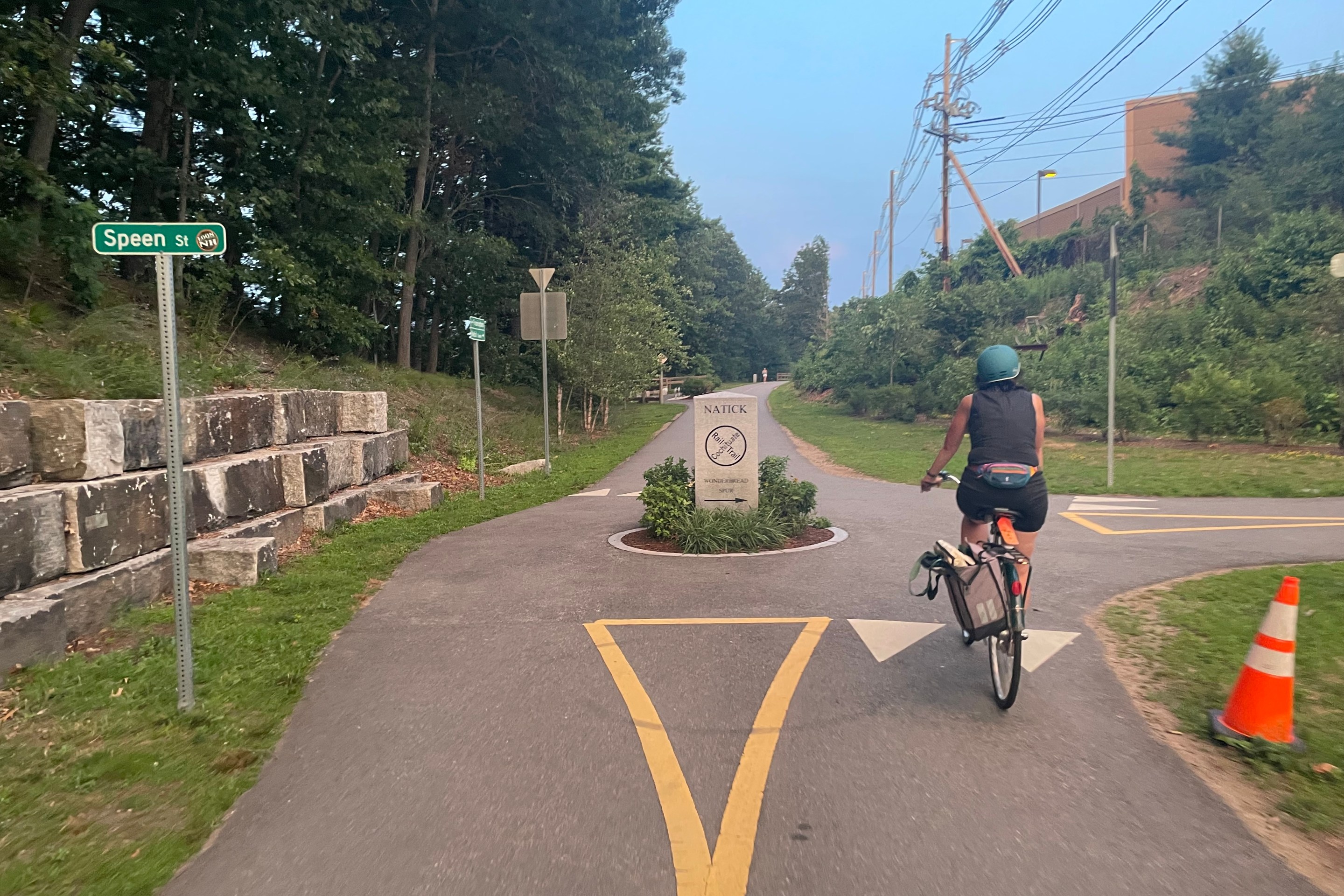 A person rides their bike along a straight, flat, paved trail lined with trees to the left and a power line at right. The rider is approaching a small traffic circle junction with a street sign at left that reads "Speen St". In the middle of the junction is a stone column with the words "Natick Cochituate Rail Trail"