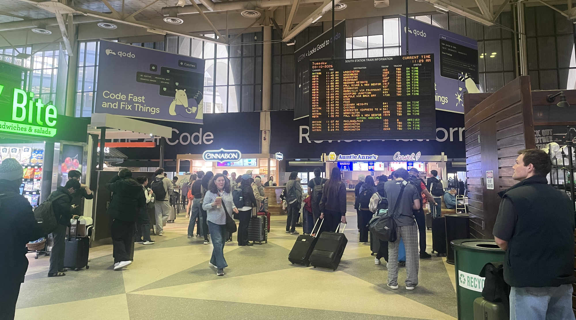 A crowded train hall filled with people, many of whom are carrying luggage. A large electronic sign hanging from the high ceiling displays upcoming train departures.