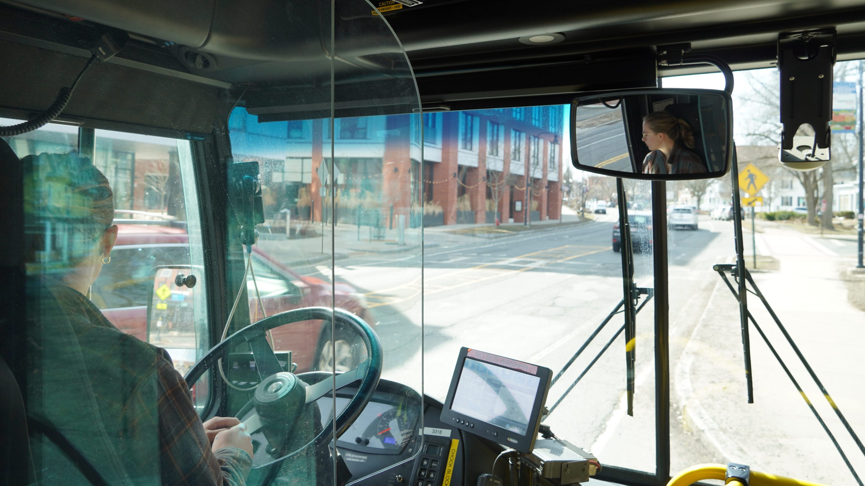 A photo of UMass Transit employee Ash Larner driving a city bus don a downtown street. The photo is taken from behind the driver's seat of a city bus, with Larner's face visible in profile the rear-view mirror at upper right.