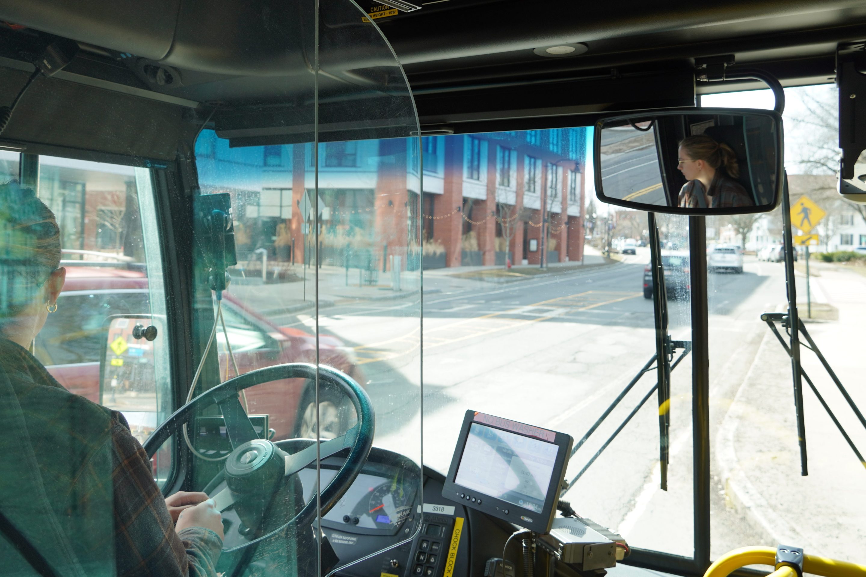 A photo of UMass Transit employee Ash Larner driving a city bus don a downtown street. The photo is taken from behind the driver's seat of a city bus, with Larner's face visible in profile the rear-view mirror at upper right.
