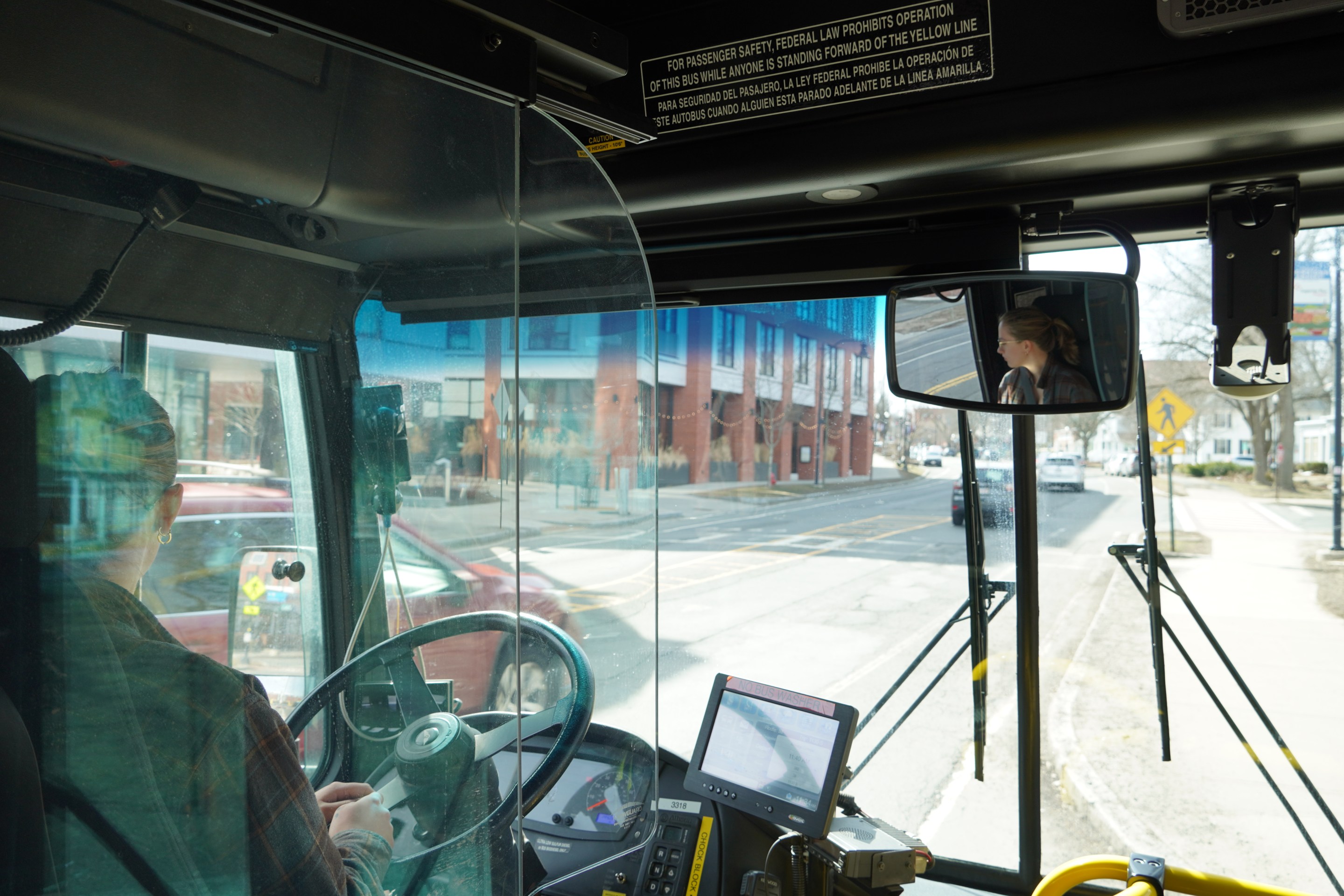 A photo of UMass Transit employee Ash Larner driving a city bus don a downtown street. The photo is taken from behind the driver's seat of a city bus, with Larner's face visible in profile the rear-view mirror at upper right.