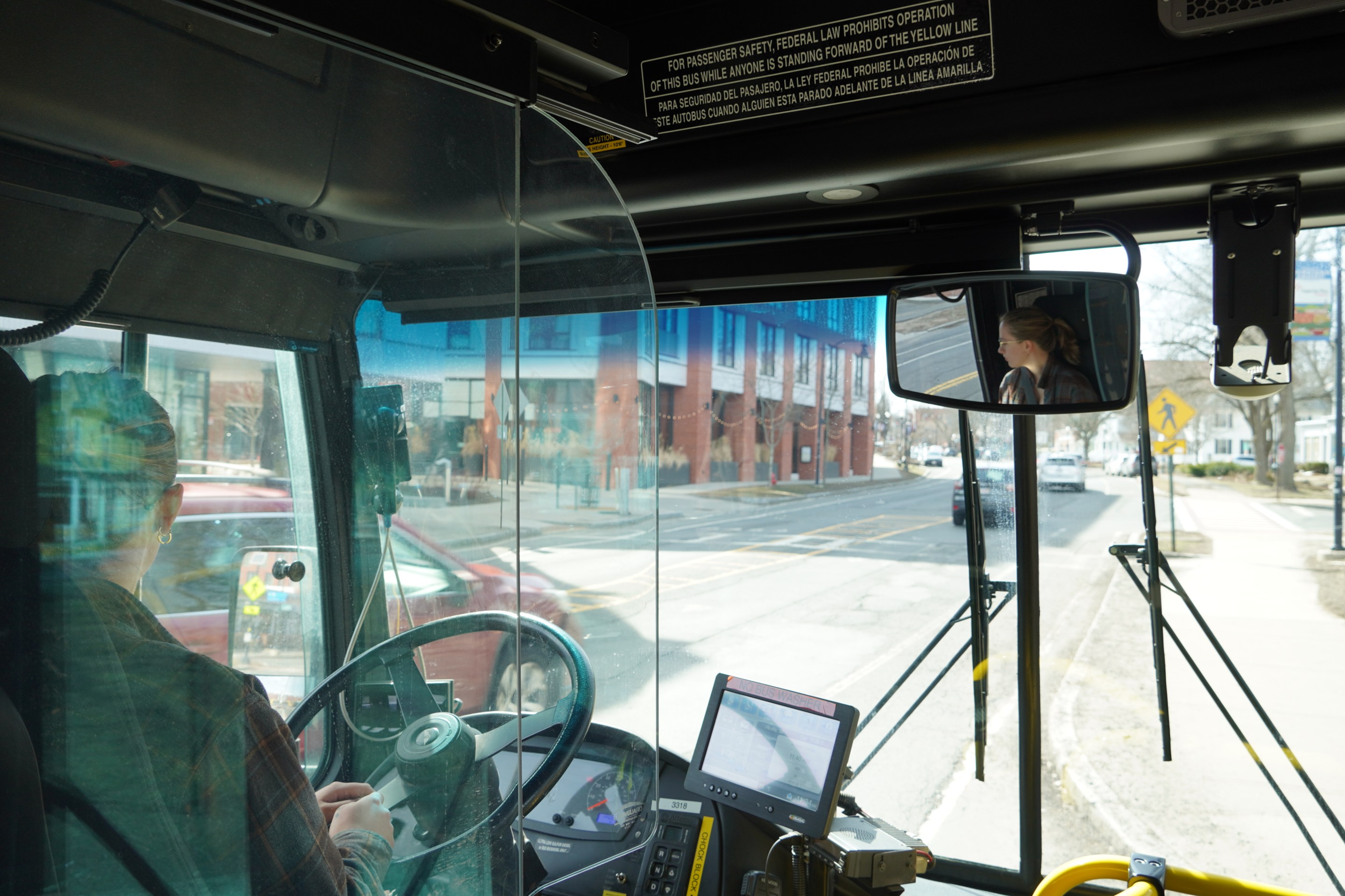 A photo of UMass Transit employee Ash Larner driving a city bus don a downtown street. The photo is taken from behind the driver's seat of a city bus, with Larner's face visible in profile the rear-view mirror at upper right.