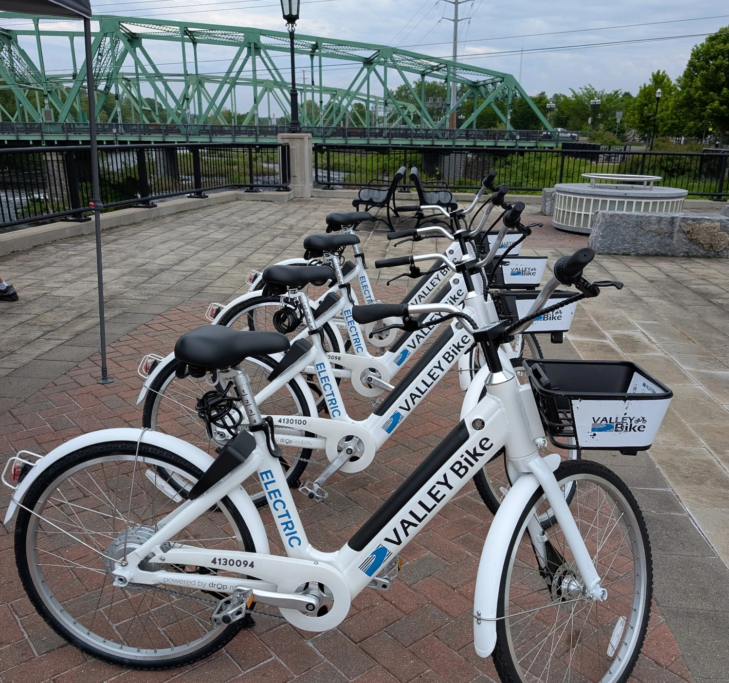A row of white electric bikes are parked next to each other on a brick plaza in front of a steel truss bridge visible in the distance. The bikes have the words "ValleyBike" on their downtubes.