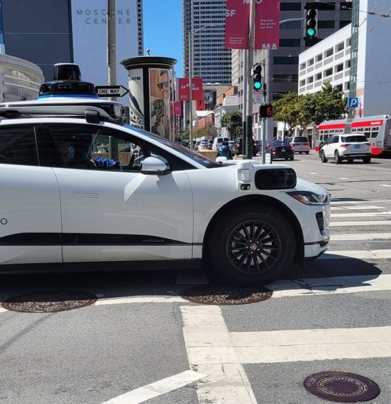 A Waymo autonomous taxi blocks a crosswalk in the middle of a large city.