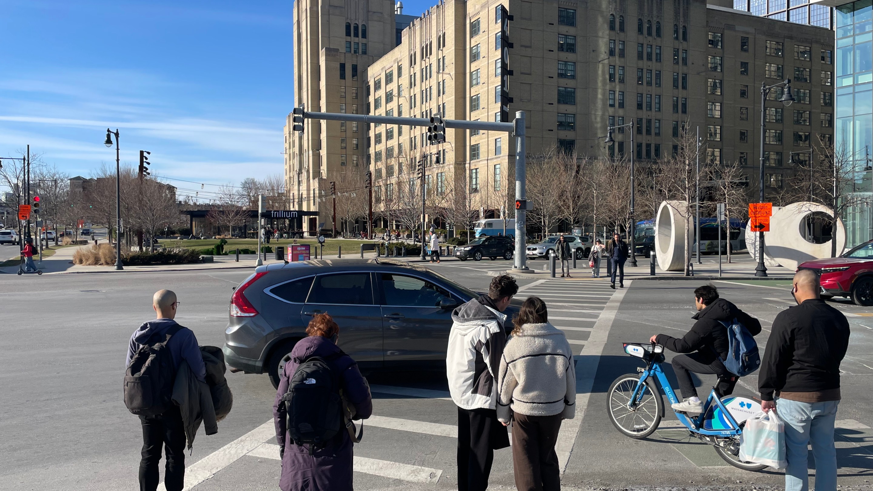 A crowd of pedestrians in winter coats waits to cross a wide intersection with a tall art deco-style warehouse building in the distance on the far side of the crosswalk.