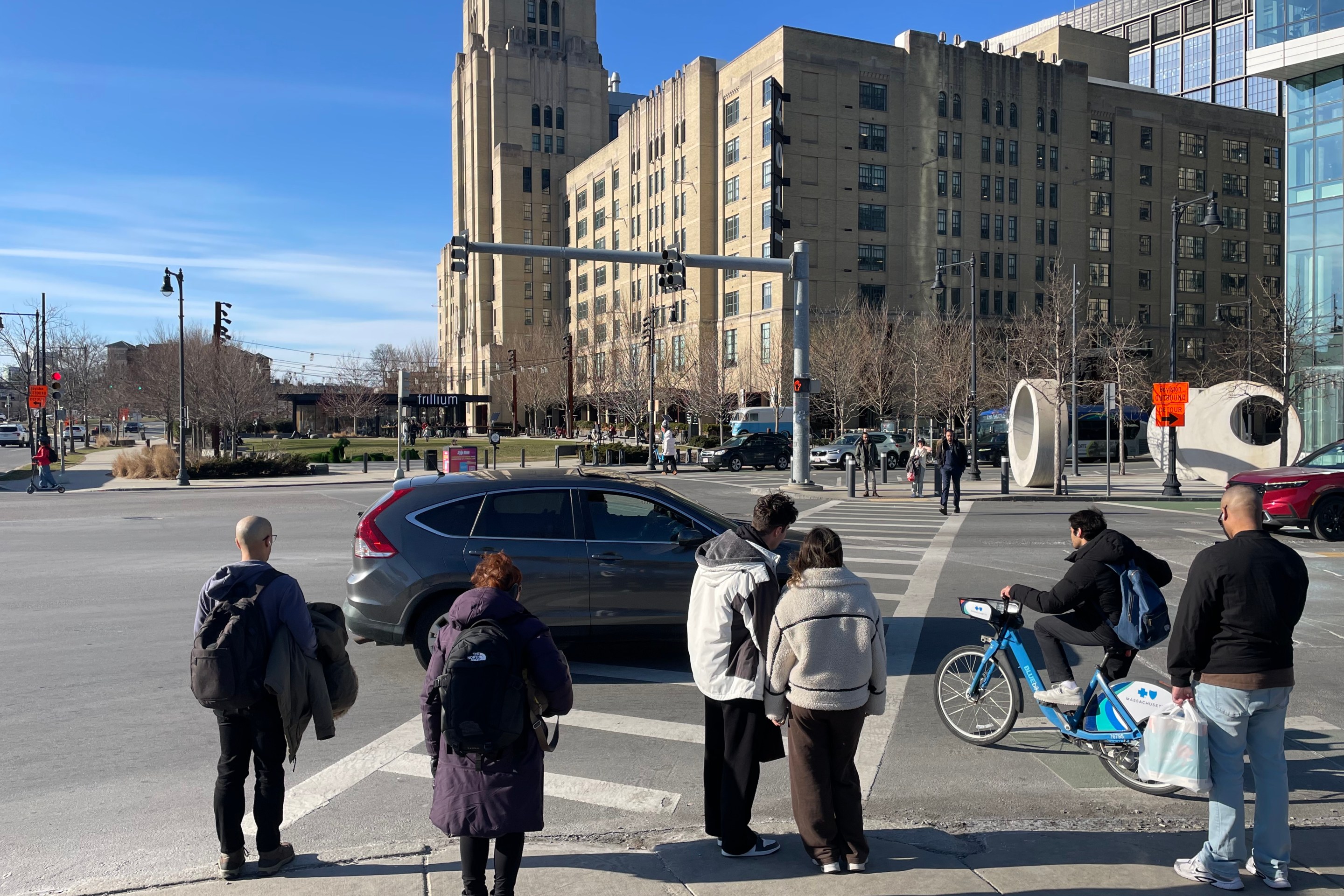A crowd of pedestrians in winter coats waits to cross a wide intersection with a tall art deco-style warehouse building in the distance on the far side of the crosswalk.